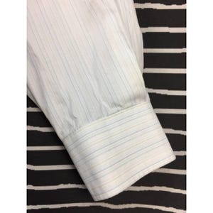 May include: A long-sleeved white dress shirt cuff featuring a subtle pinstripe pattern in light blue and tan. The fabric has a fine vertical texture, and the cuff is finished with a structured band and visible stitching along the edges of the sleeve.