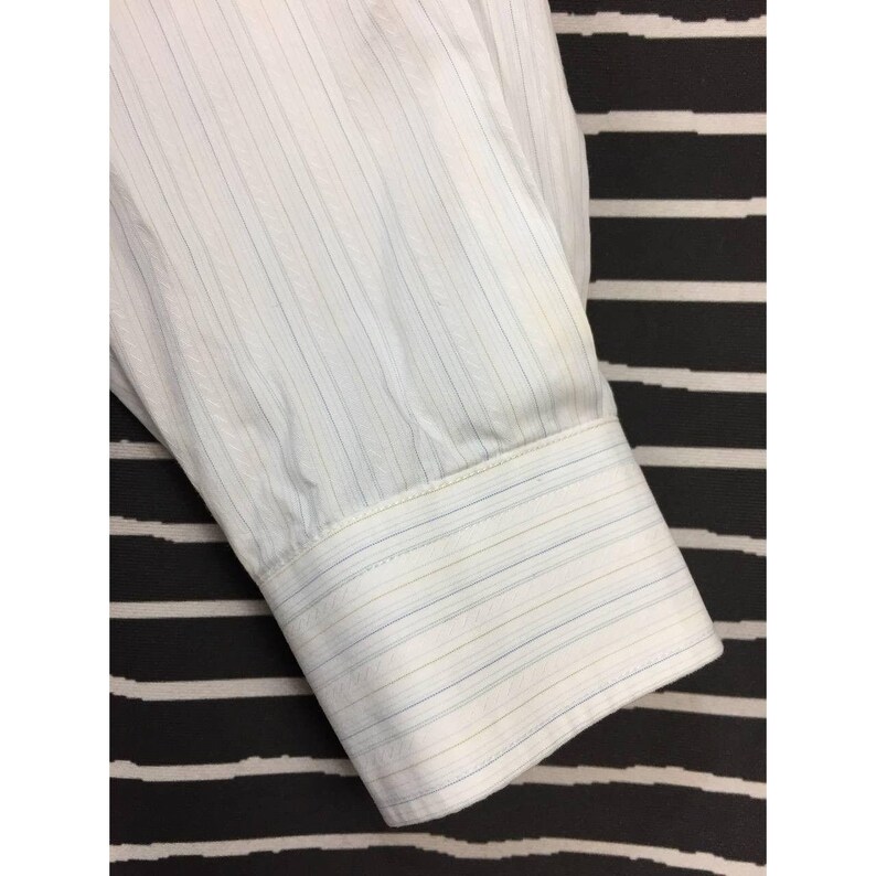 May include: A long-sleeved white dress shirt cuff featuring a subtle pinstripe pattern in light blue and tan. The fabric has a fine vertical texture, and the cuff is finished with a structured band and visible stitching along the edges of the sleeve.