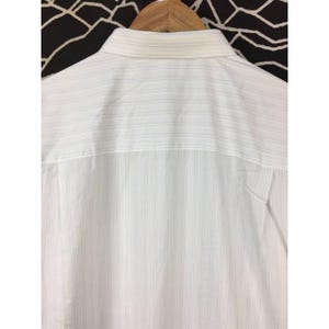 May include: A white button-down shirt featuring a subtle pattern of thin, vertical pinstripes in light blue and grey. The garment includes a structured collar and is displayed on a wooden hanger, showing the upper back portion of the shirt.
