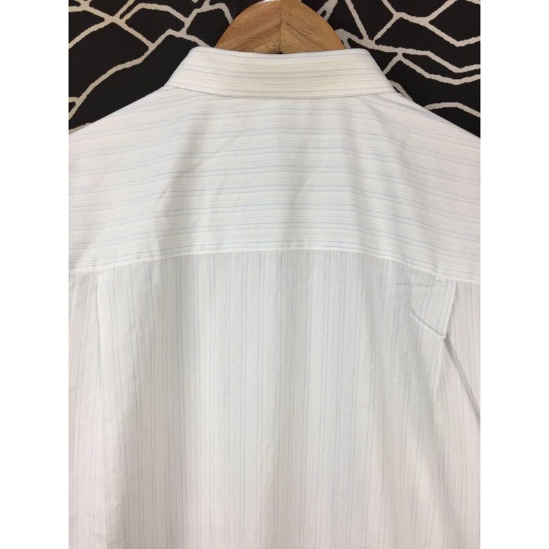 May include: A white button-down shirt featuring a subtle pattern of thin, vertical pinstripes in light blue and grey. The garment includes a structured collar and is displayed on a wooden hanger, showing the upper back portion of the shirt.