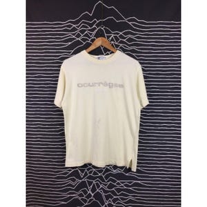 Op de afbeelding: Cr&egrave;mekleurig Courr&egrave;ges T-shirt met korte mouwen. De merknaam is in zilver op de borst gedrukt. Het shirt hangt aan een houten hanger tegen een zwart-wit patroon.