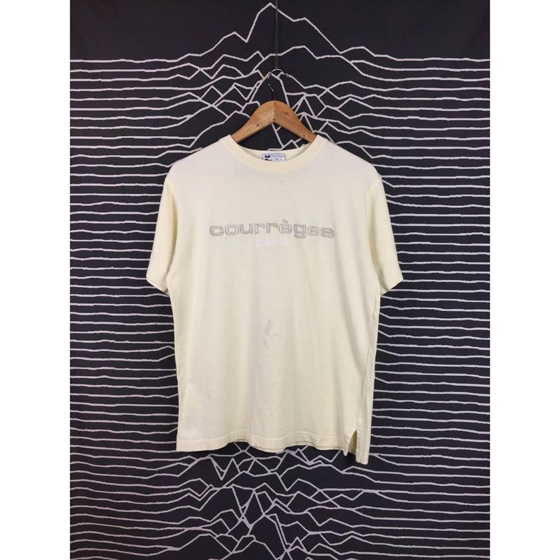 Op de afbeelding: Cr&egrave;mekleurig Courr&egrave;ges T-shirt met korte mouwen. De merknaam is in zilver op de borst gedrukt. Het shirt hangt aan een houten hanger tegen een zwart-wit patroon.
