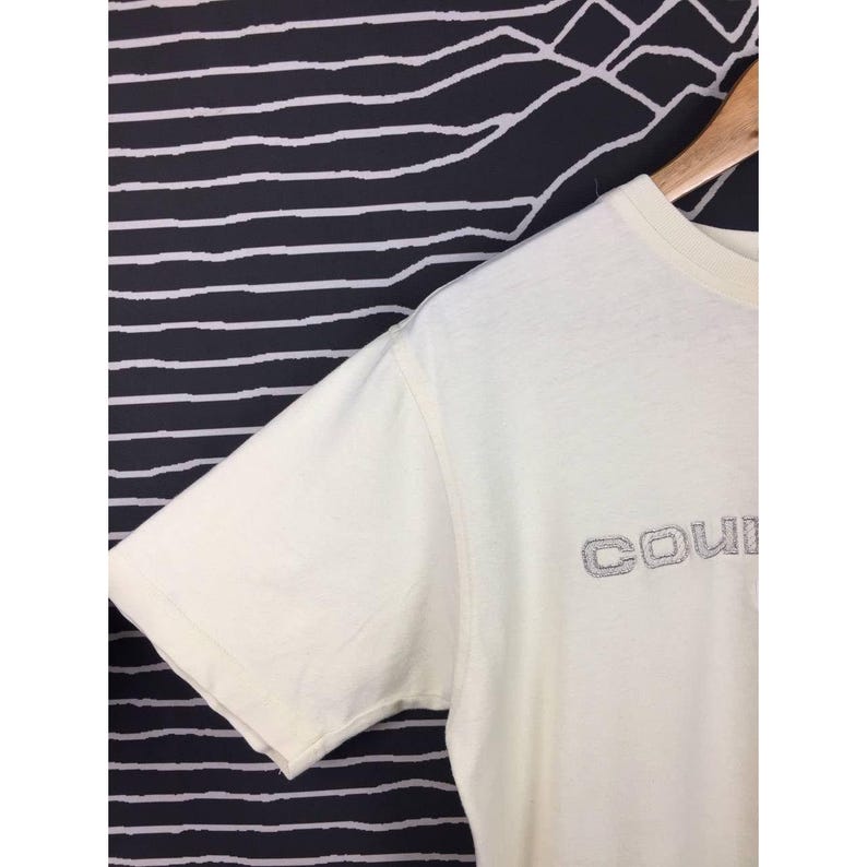 Op de afbeelding: Cr&egrave;mekleurig T-shirt met korte mouwen met het woord "COUI" geborduurd in zilvergrijs garen over de borst. Het shirt hangt aan een houten hanger tegen een zwart-wit gestreepte achtergrond.