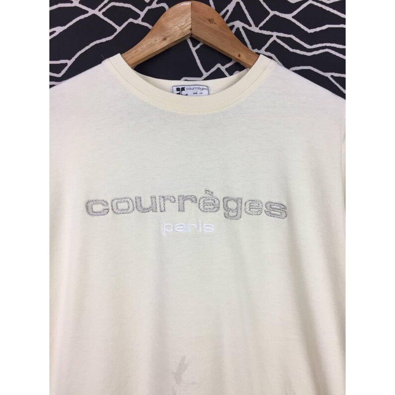 Op de afbeelding: Cr&egrave;mekleurig T-shirt met de geborduurde merknaam "courr&egrave;ges" in zilverkleurig garen. Het woord "paris" is eronder geborduurd in een kleiner lettertype. Het shirt hangt aan een houten hanger.