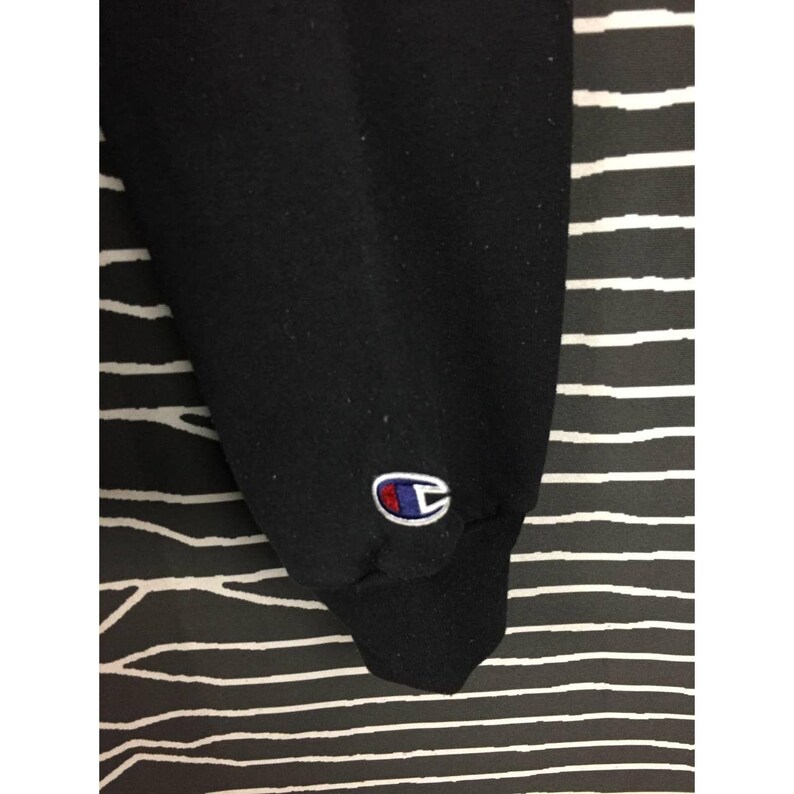 May include: Black sweatshirt sleeve with a Champion logo patch in red, white, and blue. The sleeve is made of a thick, dark fabric. The background is a black and white striped pattern.