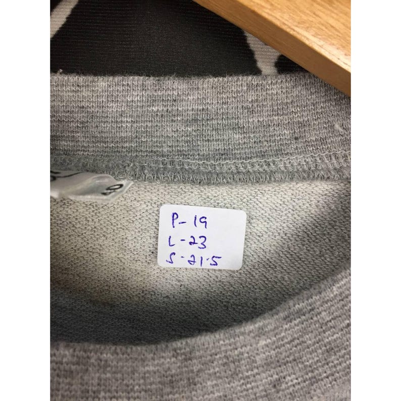 Peut inclure: Gros plan sur un col de sweat-shirt gris. Une &eacute;tiquette blanche avec du texte manuscrit noir est attach&eacute;e, affichant les chiffres P-19, L-23 et S-21.5. Le tissu a un aspect textur&eacute;.