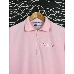 May include: A light pink polo shirt with a half-zip closure and a collared neckline. The shirt features embroidered floral and logo details. The shirt is hanging on a wooden hanger against a black and white patterned background.