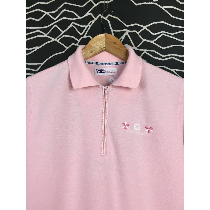 May include: A light pink polo shirt with a half-zip closure and a collared neckline. The shirt features embroidered floral and logo details. The shirt is hanging on a wooden hanger against a black and white patterned background.