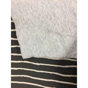 Peut inclure: Gros plan sur un tissu gris clair, possiblement un t-shirt ou un sweat-shirt, avec une surface l&eacute;g&egrave;rement textur&eacute;e. Le tissu pr&eacute;sente quelques d&eacute;colorations. Le fond est un motif &agrave; rayures noires et blanches.