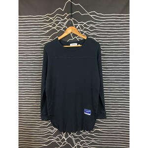 Peut inclure: Un t-shirt à manches longues bleu marine avec un col rond et un ourlet incurvé. Le t-shirt présente une couture horizontale sur la poitrine et une petite étiquette bleue et blanche en bas à gauche. Suspendu à un cintre en bois.