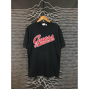 Könnte beinhalten: Schwarzes Guess T-Shirt mit einem rot-weißen Logo mit der Aufschrift "Guess Since 1981". Das T-Shirt hängt an einem Holzbügel vor einem schwarz-weiß gemusterten Hintergrund. Das Shirt hat kurze Ärmel.