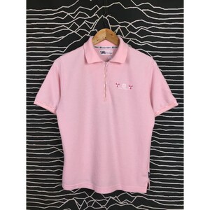 May include: A light pink polo shirt with short sleeves and a zipper closure. The shirt has a collar and a small embroidered design near the zipper. The shirt is hanging on a wooden hanger against a black and white patterned background.