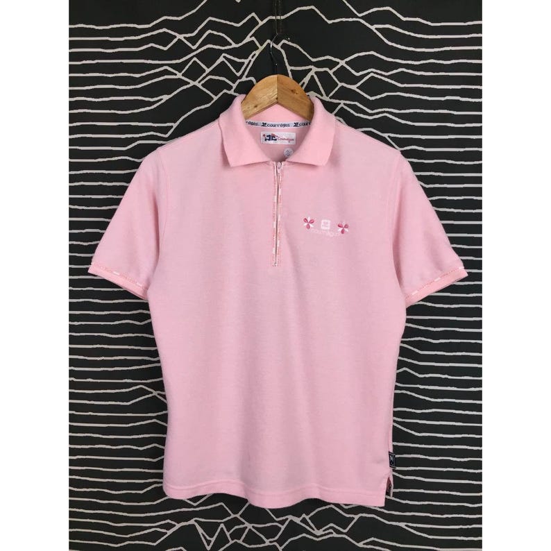 May include: A light pink polo shirt with short sleeves and a zipper closure. The shirt has a collar and a small embroidered design near the zipper. The shirt is hanging on a wooden hanger against a black and white patterned background.