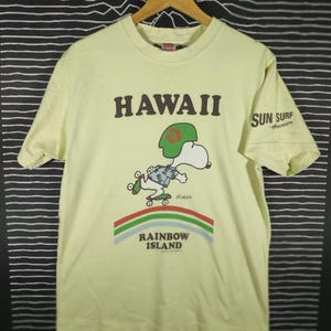 May include: Light yellow t-shirt with the word "HAWAII" in brown block letters. A cartoon character wearing a green hat and Hawaiian shirt is skateboarding. The shirt also features the words "SUN SURF" and "RAINBOW ISLAND".