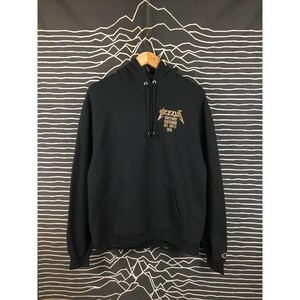 May include: Black hoodie with a front kangaroo pocket and a drawstring hood. The front features a gold graphic with the word "Yeezy" and other text. The Champion logo is visible on the left sleeve. The hoodie is hanging on a wooden hanger.