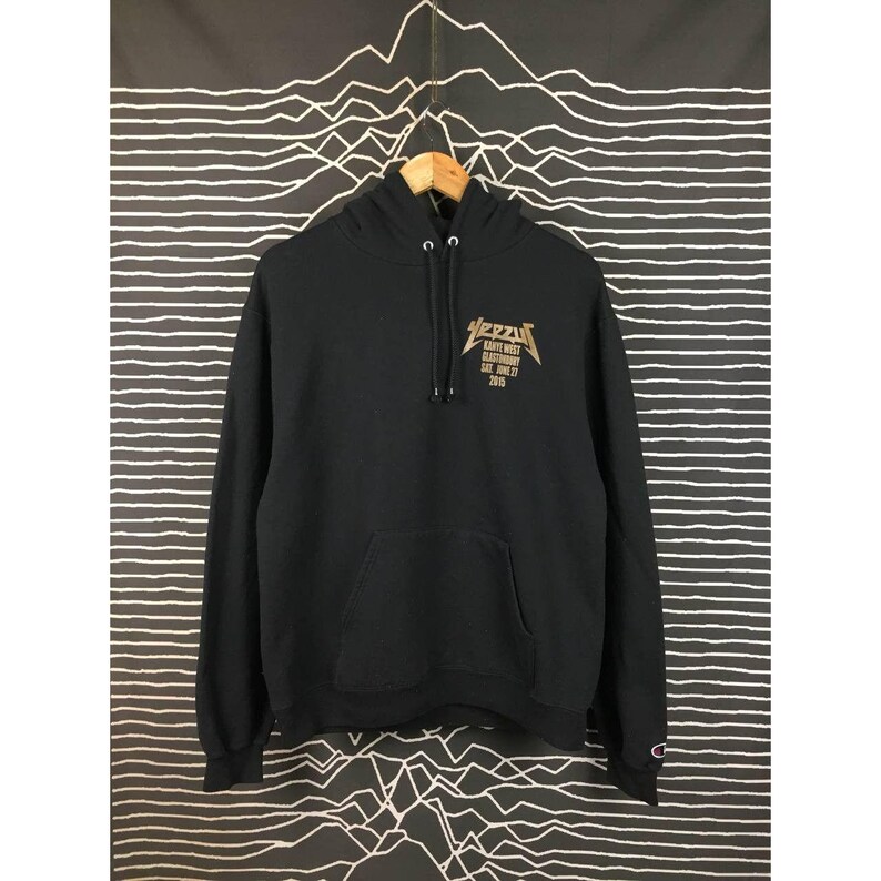May include: Black hoodie with a front kangaroo pocket and a drawstring hood. The front features a gold graphic with the word "Yeezy" and other text. The Champion logo is visible on the left sleeve. The hoodie is hanging on a wooden hanger.