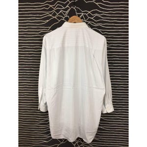 May include: A long-sleeve button-down shirt in a light white color with a subtle vertical pinstripe pattern. The shirt features a classic collar, a single chest pocket, and buttoned cuffs, displayed hanging on a wooden hanger against a dark background.