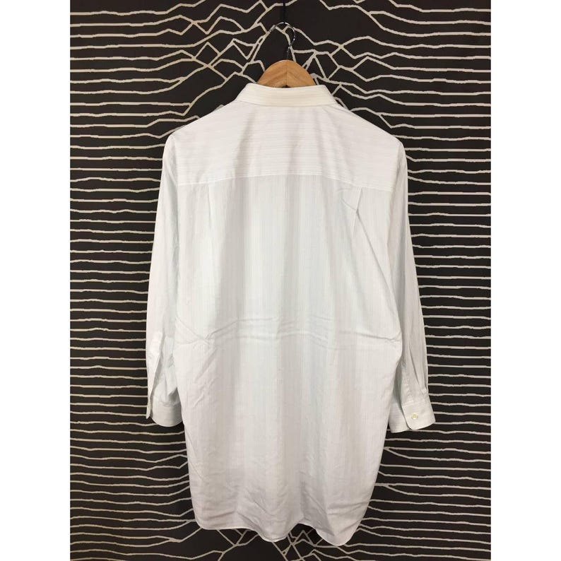 May include: A long-sleeve button-down shirt in a light white color with a subtle vertical pinstripe pattern. The shirt features a classic collar, a single chest pocket, and buttoned cuffs, displayed hanging on a wooden hanger against a dark background.