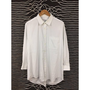 May include: A long-sleeve button-down shirt in white with a subtle vertical pinstripe pattern. The shirt features a classic collar, a single chest pocket on the left side, and a buttoned front placket with matching white buttons throughout the garment.