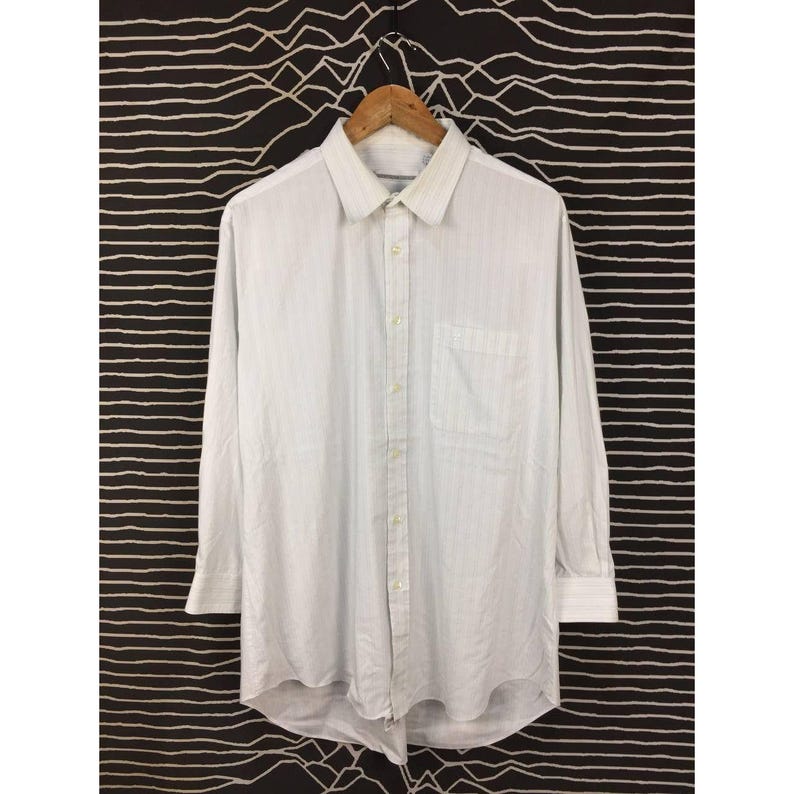 May include: A long-sleeve button-down shirt in white with a subtle vertical pinstripe pattern. The shirt features a classic collar, a single chest pocket on the left side, and a buttoned front placket with matching white buttons throughout the garment.