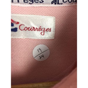 May include: Close-up of a pink fabric with a label that reads "Courr&egrave;ges" in red script. The label has a red and blue design. A white sticker with the numbers "19" and "24" is also visible. The fabric has a textured appearance.