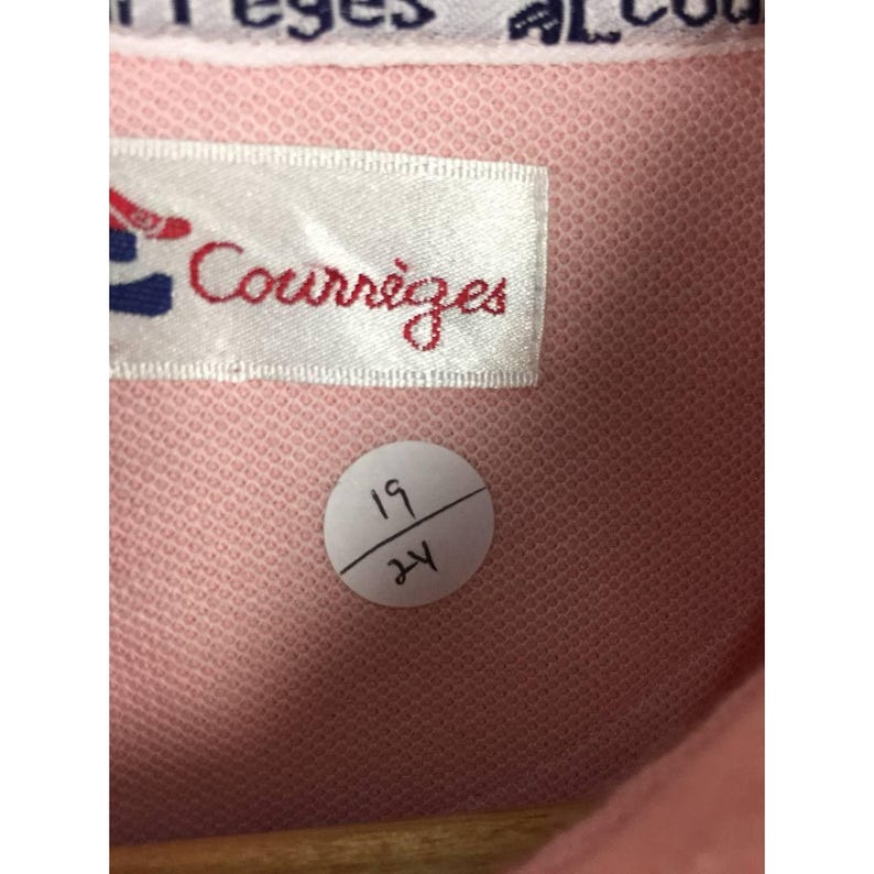 May include: Close-up of a pink fabric with a label that reads "Courr&egrave;ges" in red script. The label has a red and blue design. A white sticker with the numbers "19" and "24" is also visible. The fabric has a textured appearance.