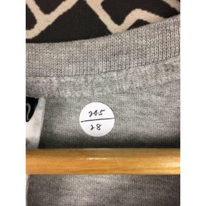 Peut inclure: Gros plan sur un col de t-shirt gris avec une &eacute;tiquette blanche et un autocollant rond. L'autocollant porte les chiffres "205" sur "28" &agrave; l'encre noire. Le t-shirt est sur un cintre en bois.
