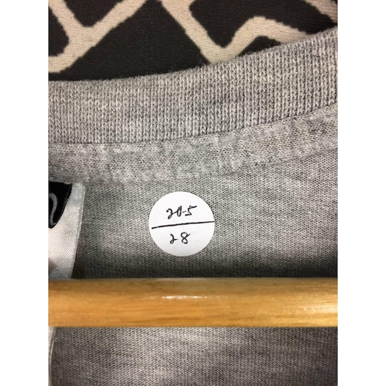 Peut inclure: Gros plan sur un col de t-shirt gris avec une &eacute;tiquette blanche et un autocollant rond. L'autocollant porte les chiffres "205" sur "28" &agrave; l'encre noire. Le t-shirt est sur un cintre en bois.