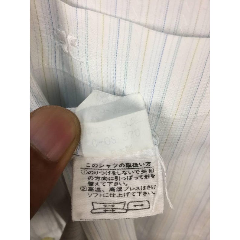 May include: A white dress shirt featuring thin vertical pinstripes in light blue and yellow. The garment includes a white fabric care label with Japanese text and a small embroidered logo on the chest pocket area, visible near the collar.
