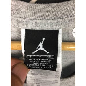 Peut inclure: Gros plan sur une &eacute;tiquette de t-shirt grise avec le logo Jumpman blanc. L'&eacute;tiquette indique "MADE IN HONDURAS OF U.S.A. FABRICS FABRIQUE AU HONDURAS AVEC TISSU PROVENANT DES E.U."