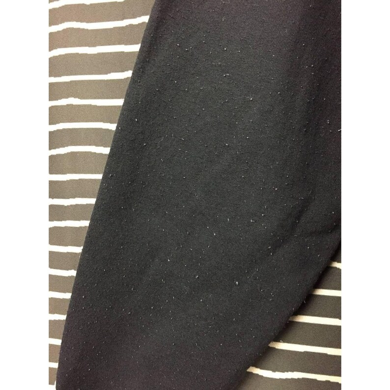May include: Close-up of a black fabric with a slightly textured surface. The fabric is covered in small, light-colored particles. The background features a striped pattern in shades of brown and white.