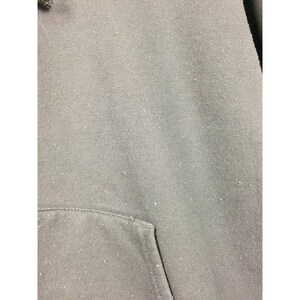 May include: Close-up of a gray hoodie with a kangaroo pocket. The fabric has a slightly worn appearance with small white specks scattered across the surface. The hood is visible at the top left.