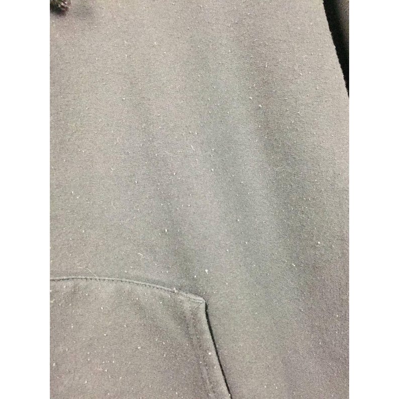 May include: Close-up of a gray hoodie with a kangaroo pocket. The fabric has a slightly worn appearance with small white specks scattered across the surface. The hood is visible at the top left.