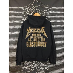 May include: Black hoodie with gold lettering featuring the word "YEEZUS" and "Kanye West" with event details. The hoodie has a drawstring hood and a kangaroo pocket. The text includes "Worthy Farm, Pilton, Somerset, SAT. JUNE 27 2015, GLASTONBURY."
