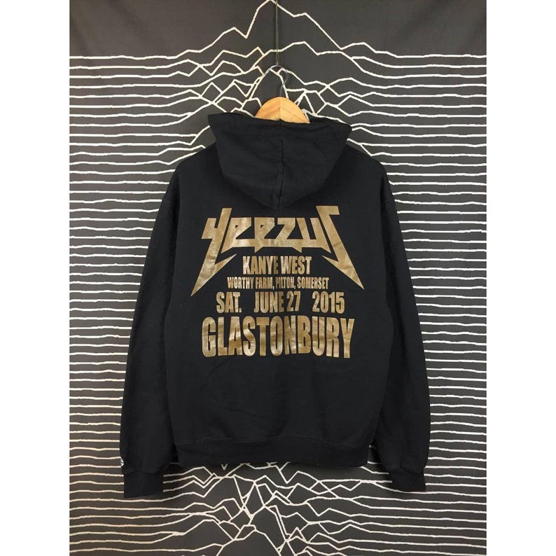 May include: Black hoodie with gold lettering featuring the word "YEEZUS" and "Kanye West" with event details. The hoodie has a drawstring hood and a kangaroo pocket. The text includes "Worthy Farm, Pilton, Somerset, SAT. JUNE 27 2015, GLASTONBURY."