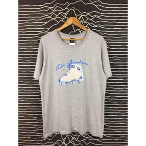 Peut inclure: T-shirt gris avec un motif bleu et blanc d'une chaussure de basket-ball et l'inscription "Air Jordan" en bleu. Le t-shirt a un col rond et des manches courtes.