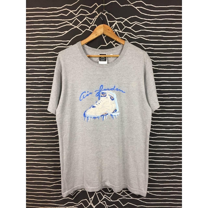 Peut inclure: T-shirt gris avec un motif bleu et blanc d'une chaussure de basket-ball et l'inscription "Air Jordan" en bleu. Le t-shirt a un col rond et des manches courtes.