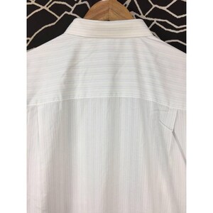 May include: A white button-down shirt featuring a subtle pattern of thin, vertical pinstripes in light blue and grey. The garment includes a structured collar and is displayed on a wooden hanger, showing the upper back portion of the shirt.
