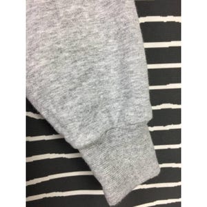 Peut inclure: Gros plan sur une manche de sweat-shirt grise. Le poignet est c&ocirc;tel&eacute; et le tissu a une texture chin&eacute;e. Le fond est un motif &agrave; rayures noires et blanches.