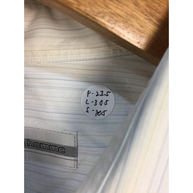 May include: A white button-down shirt featuring thin blue and tan pinstripes and a subtle diagonal weave pattern. A white circular sticker is attached to the fabric with the handwritten text P-23.5, L-30.5, S-20.5. A label reads homme.