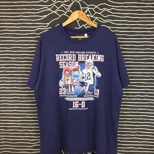 May include: A navy blue t-shirt with a graphic of Tom Brady and a teammate from the 2007 New England Patriots season. The text on the shirt reads "2007 New England Patriots * Record Breaking Season * 29 Receiving Touchdowns * 50 Passing Touchdowns * Record 589 Total Points Scored * 16-0 Undefeated Regular Season *"