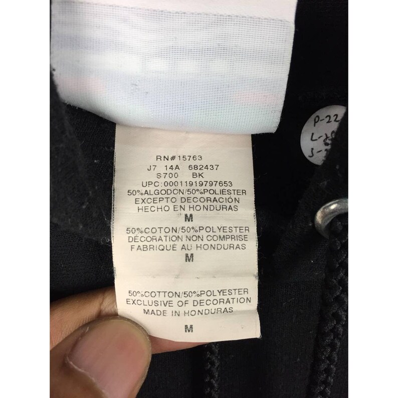 May include: Close-up of a black garment tag with text including "RN#15763", "S700 BK", and "Made in Honduras". The tag also lists fabric composition and size "M".
