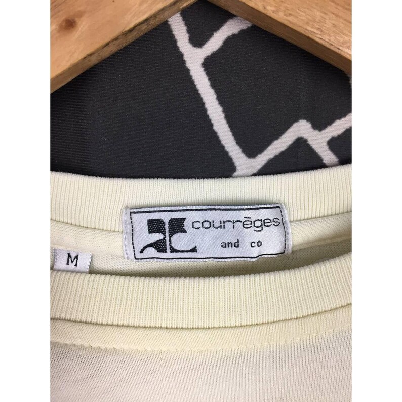 Op de afbeelding: Close-up van een cr&egrave;mekleurig t-shirt met het label "courr&egrave;ges and co." Het label is rechthoekig met zwarte tekst en een logo. Het shirt hangt aan een houten hanger. De maat "M" is zichtbaar.