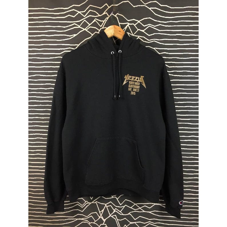 May include: Black hoodie with a front pocket and a drawstring hood. The gold text on the front reads "YEEZY" above "KANYE WEST GLASTONBURY SAT. JUNE 27 2015". The hoodie has the Champion logo on the left sleeve.