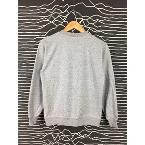 Peut inclure: Un sweat-shirt gris chin&eacute; &agrave; col rond et manches longues, suspendu &agrave; un cintre en bois. Le fond pr&eacute;sente un motif abstrait noir et blanc.