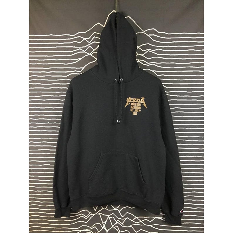 May include: Black hoodie with a front kangaroo pocket and drawstring hood. The gold text on the front reads "Yeezy" above "Kanye West Glastonbury" and "Sat. June 27 2015". The hoodie is made of a soft material.