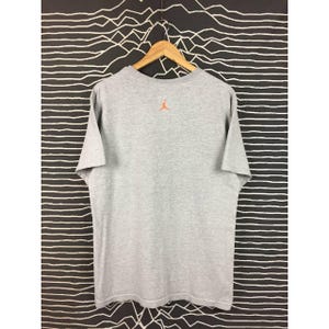 Peut inclure: Un t-shirt gris chin&eacute; &agrave; manches courtes, suspendu &agrave; un cintre en bois. Le t-shirt pr&eacute;sente un petit logo Jumpman orange dans le haut du dos. Le fond est un motif &agrave; rayures noires et blanches.