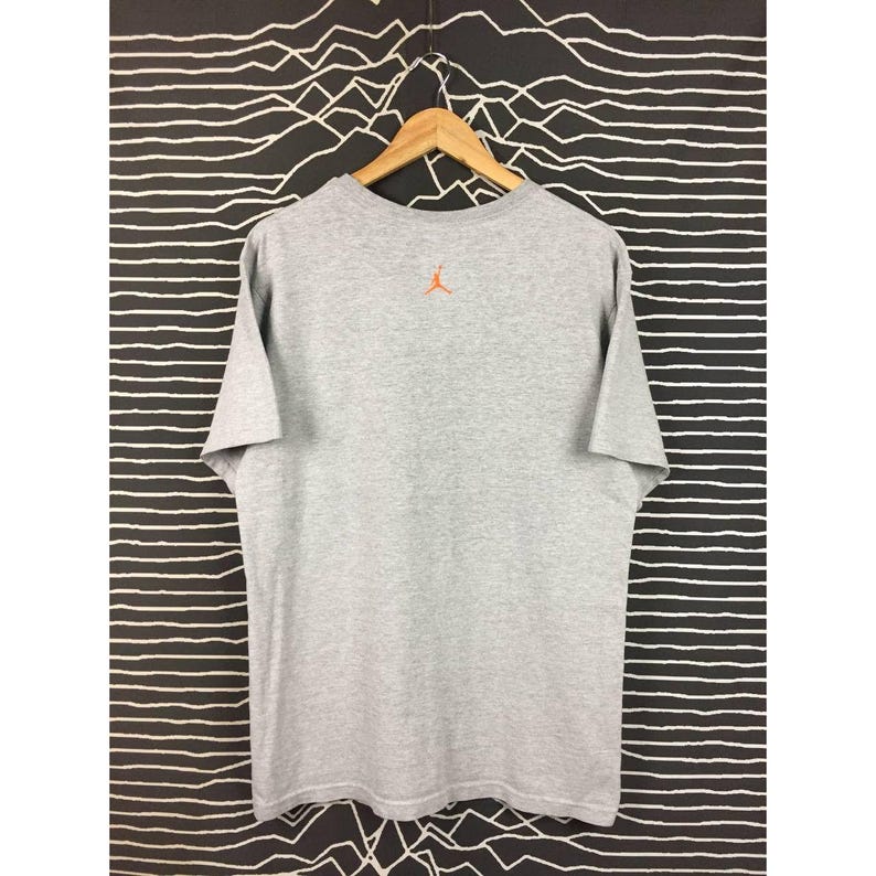 Peut inclure: Un t-shirt gris chin&eacute; &agrave; manches courtes, suspendu &agrave; un cintre en bois. Le t-shirt pr&eacute;sente un petit logo Jumpman orange dans le haut du dos. Le fond est un motif &agrave; rayures noires et blanches.