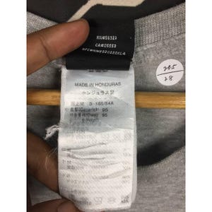 Peut inclure: Gros plan sur une &eacute;tiquette de t-shirt grise avec du texte. L'&eacute;tiquette comprend le texte "RN#56323", "CA#05553" et "RFC#NME920220KLA". L'&eacute;tiquette indique &eacute;galement "MADE IN HONDURAS" et contient du texte japonais. Un autocollant blanc avec les chiffres "20-5" et "28" est &eacute;galement visible.