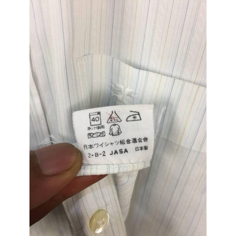 May include: A white button-down shirt featuring thin vertical stripes in light blue and yellow. A white care label is attached to the interior, displaying laundry symbols, Japanese text, the acronym JASA, and the phrase 日本製 indicating made in Japan.